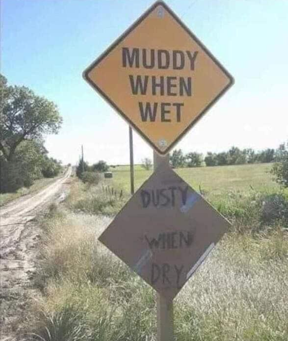 تقرأ علامة الطريق على طريق ريفي "Muddy عندما تكون رطبًا" و "Dusty عندما تجف" ، مع تسليط الضوء على الظروف المختلفة بناءً على الطقس