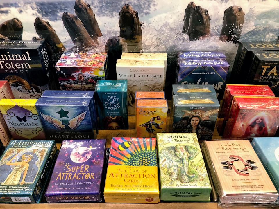 الرف يعرض مختلف طوابق بطاقة Oracle و Tarot مع تصميمات ملونة ، بما في ذلك "Light Seers Tarot" و "Law of Attraction Cards".