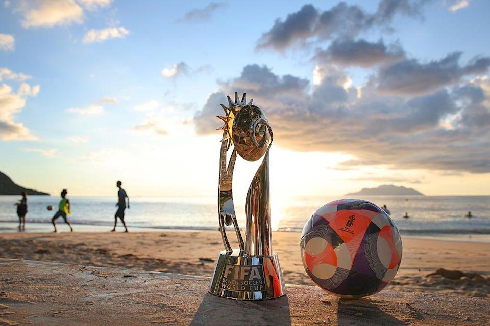 منظر مفصل للكأس وكرة المباراة الرسمية حيث يلعب الناس كرة القدم في Beau Vallon Beach. الشمس منخفضة هي السماء فوق البحر في الخلفية.