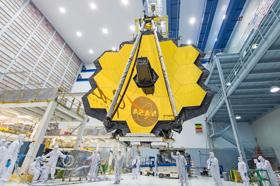 مرآة JWST ، التي تبدو وكأنها سطح لامع سداسي كبير تقريبًا يتكون من 18 سداسيًا أصغر ، يجلس في منشأة. تعكس المرآة شعار ناسا اللحم.
