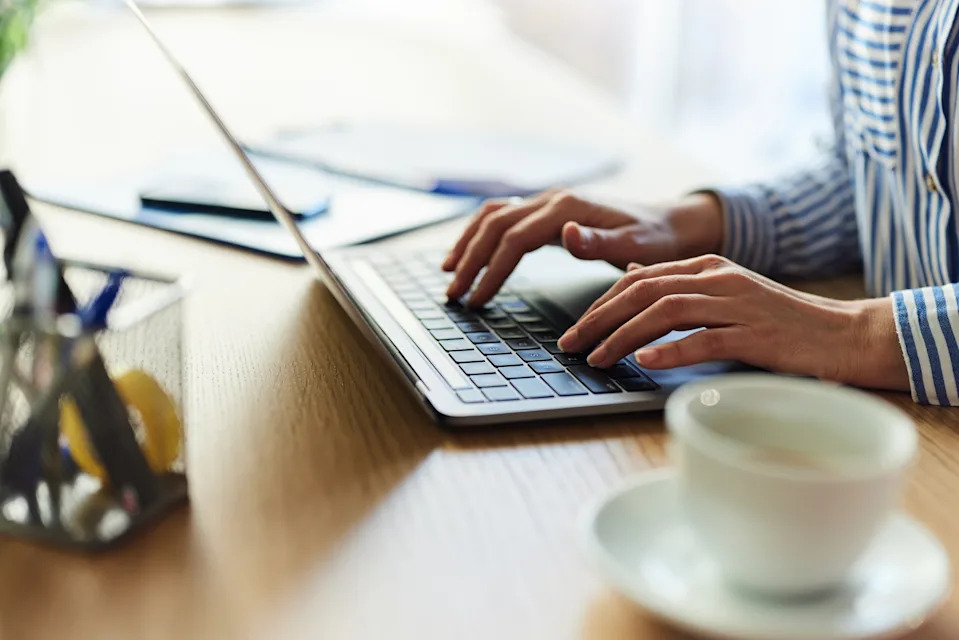 شخص يكتب على جهاز كمبيوتر محمول على مكتب خشبي مع فنجان قهوة قريب ، يرتدي قميصًا مخططًا.