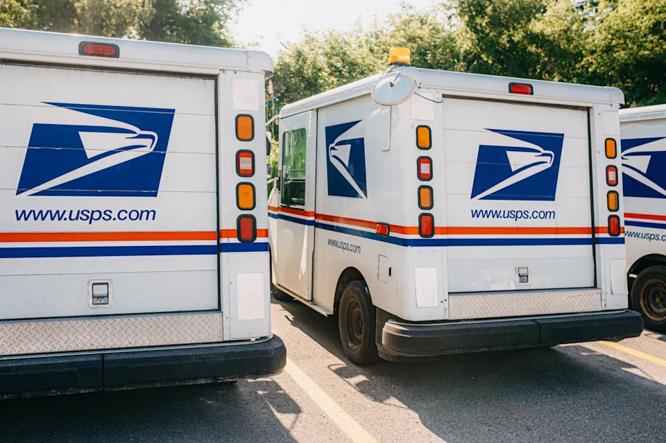 ثلاث شاحنات توصيل USPS متوقفة ، تعرض شعارات الخدمة البريدية وعناوين موقع الويب.