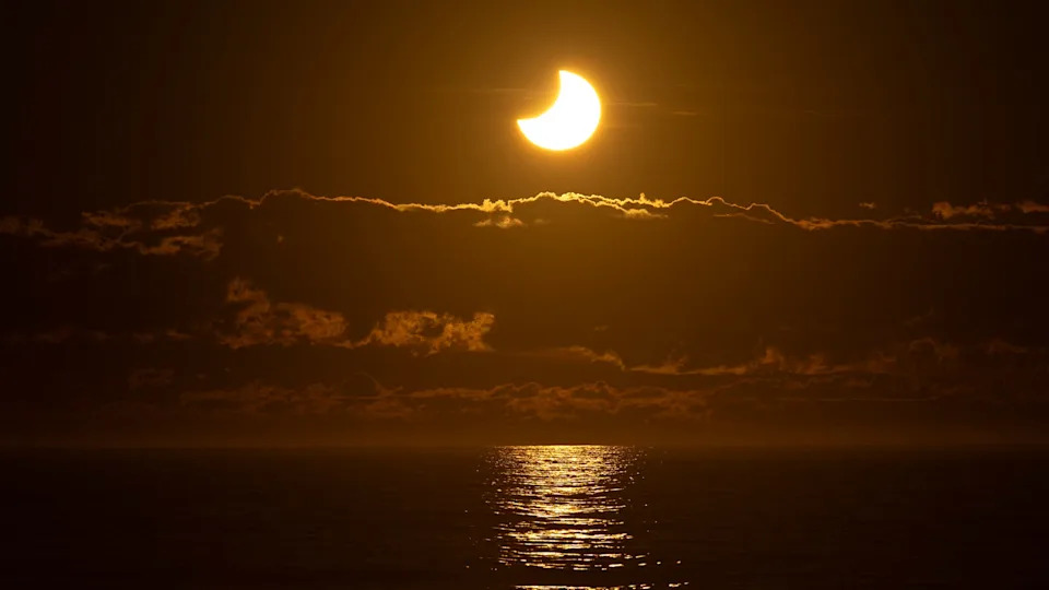 يتم تصوير قرص الشمس خلال كسوف جزئي شمسي مع الجانب الأيسر العلوي مخبأ بواسطة صورة ظلية منحنية للقمر على سماء داكنة بألوان برتقالية. يتم وضع الشمس فوق ضفة سحابية ، مع المحيط أدناه مع ضوءها.