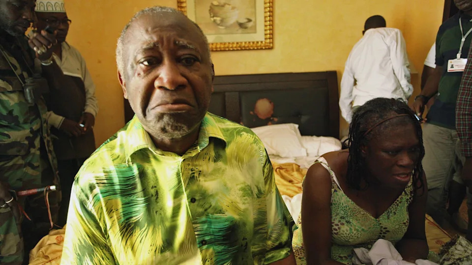 يجلس Laurent Gbagbo و Simone Gbagbo على حافة السرير ، مع تعبيرات Crestfall على وجوههما. الشخصيات ، بما في ذلك رجل يرتدي زيًا عسكريًا ، تقف وراءها.