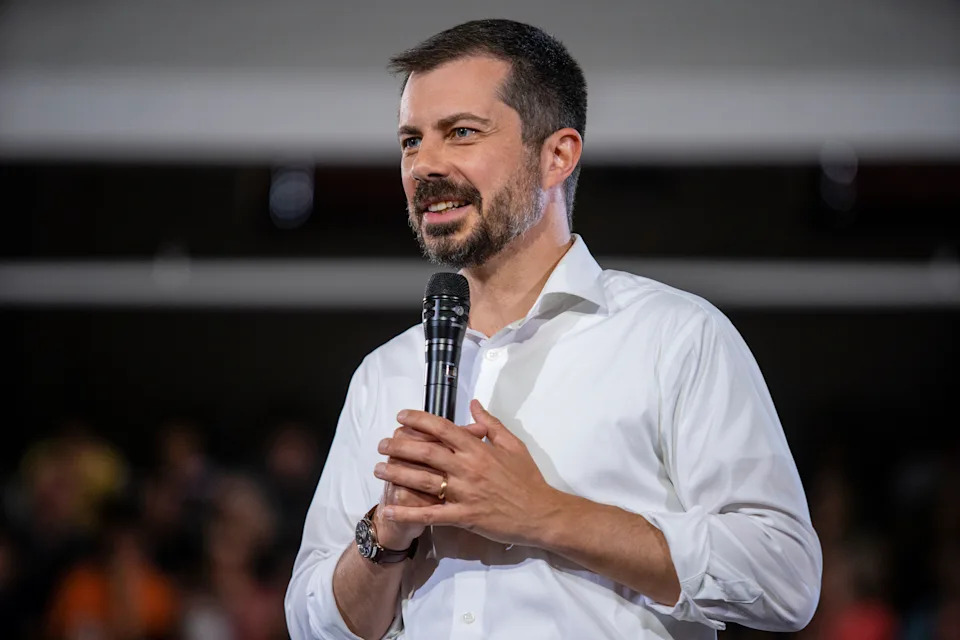 شخص يرتدي قميصًا أبيض يحمل ميكروفونًا يتحدث إلى جمهور في حدث ما. يبدو أن الإعداد بمثابة مشاركة في التحدث أمام الجمهور