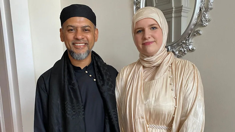 محمد موكوزال علي مع ابنته عاليه نايت. محمد يرتدي اللون الأسود بالكامل. عاليه ترتدي حجابًا وفستانًا من الحرير الكريمي. يقفون جنبًا إلى جنب وينظرون إلى الكاميرا ويبتسمون.