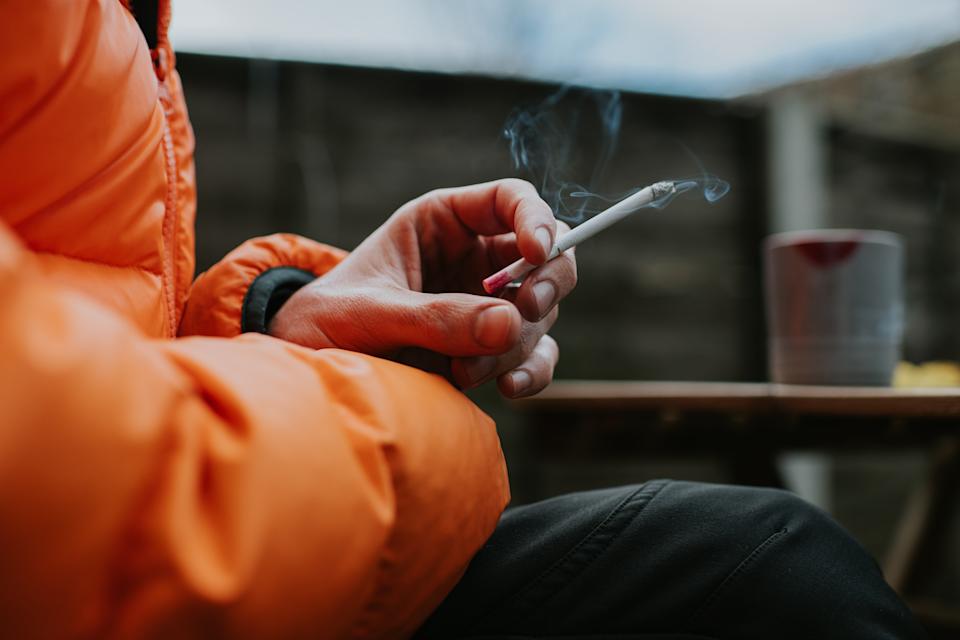 شخص يجلس ويحمل سيجارة مشتعلة في الخارج ويرتدي سترة منتفخة. تظهر طاولة بها كوب في الخلفية