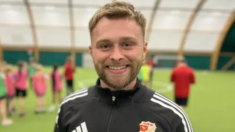 آندي تاي يبتسم ويرتدي قميصًا أسودًا من Swindon Town FC Foundation. لديه شعر قصير بني اللون ولحية.