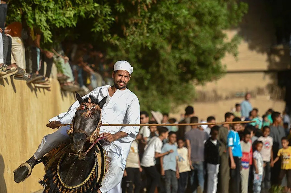 رجل يلوح بهراوة خشبية على ظهور الخيل. حشد من خلفه يراقب.