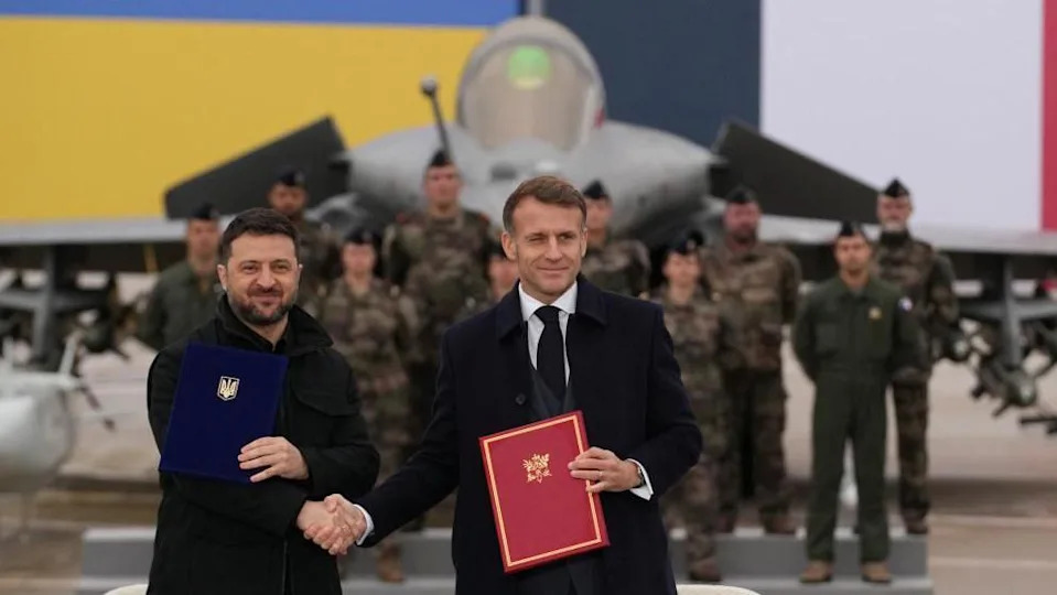 رجلان يقفان مبتسمين أمام طائرة مقاتلة في حظيرة الطائرات.