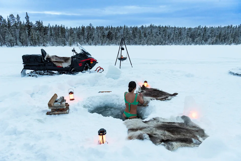 المنظر الخلفي لفتاة جميلة أثناء الاستحمام بالجليد في حفرة مفتوحة على بحيرة متجمدة محاطة بغابة مغطاة بالثلوج، Jokkmokk، السويد، الدول الاسكندنافية