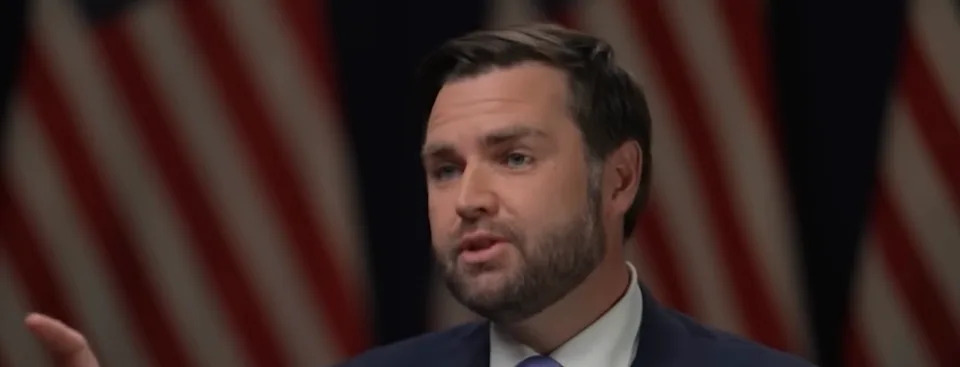 شخص يرتدي بدلة يتحدث، وأعلام الولايات المتحدة غير واضحة في الخلفية