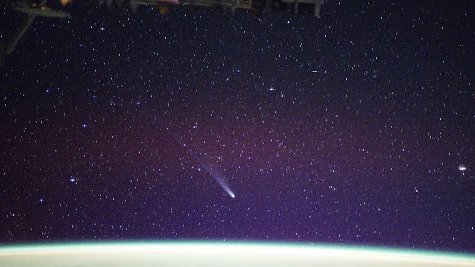 يظهر خط من اللون الأبيض أثناء تحرك المذنب ليمون نحو أسفل يمين الصورة في سماء الليل الأرجوانية المرصعة بالنجوم. يوجد في الأسفل أضواء منحنية باللونين الأبيض والأخضر