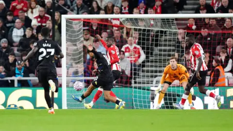 PA Media إليمان ندياي يرتدي طقمًا أسود. ويمكن رؤيته وهو يركل الكرة في المرمى في ملعب كرة قدم بينما يحاول لاعب يرتدي القمصان الحمراء والبيضاء اعتراضها. إنهم في ملعب كرة قدم أخضر ويرتدي حارس المرمى قميصًا برتقاليًا. 