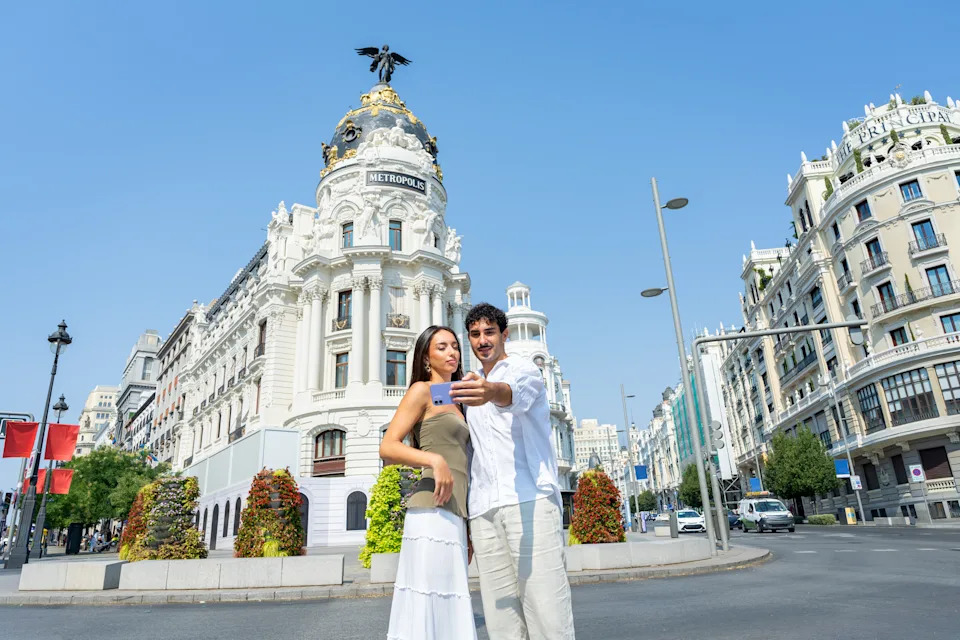 زوجان شابان يقضيان إجازة يبتسمان ويحملان هاتفًا ذكيًا ويلتقطان صورة ذاتية أمام مبنى متروبوليس التاريخي في مدريد، ويتشاركان تجربة السفر. زوجان شابان يلتقطان صورة شخصية أمام مبنى متروبوليس الشهير في شارع غران فيا، ويلتقطان ذكريات سفر مبهجة خلال إجازتهما. يخلق اليوم المشمس والهندسة المعمارية الأوروبية الأنيقة خلفية مثالية للسياحة الحديثة والرومانسية. تعكس هذه الصورة الحب والتواصل والمغامرة، وهي مثالية لتوضيح مفاهيم نمط الحياة الرقمي والتصوير الفوتوغرافي أثناء السفر ومشاهدة معالم المدينة والأزواج الذين يستمتعون بالعطلات الحضرية معًا. مدريد، اسبانيا