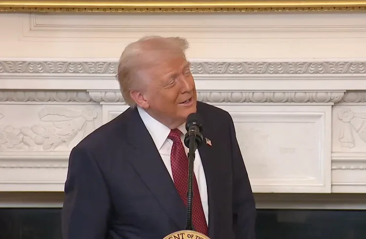 شخص يرتدي بدلة وربطة عنق يتحدث على منصة في الداخل مع وجود مدفأة مزخرفة في الخلفية