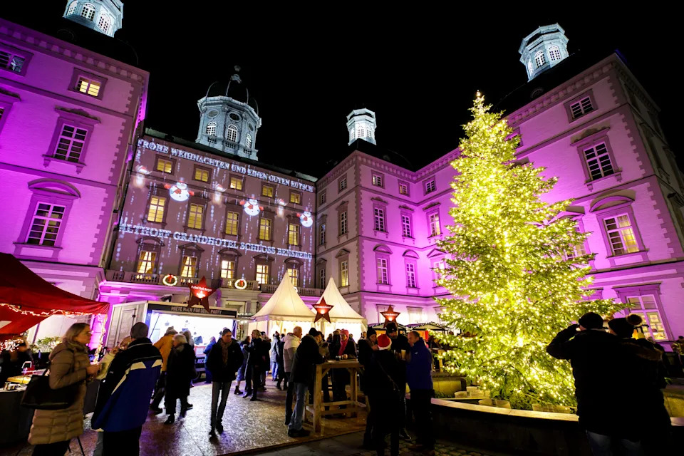 الواجهة المضيئة لفندق Althoff Grandhotel Schloss Bensberg مع شجرة عيد الميلاد المتوهجة والزوار يستمتعون بالسوق الاحتفالي ليلاً.