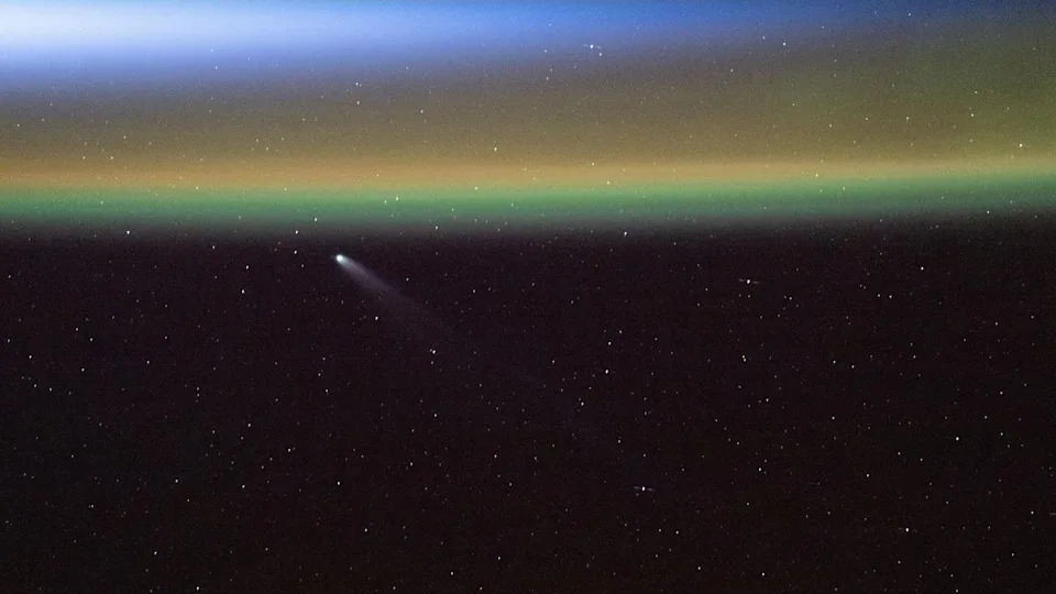 يظهر خط أبيض مقابل ظلام الفضاء المذنب سوان وهو يتحرك نحو أعلى يسار الصورة بالقرب من سلسلة من الخطوط الصفراء والخضراء والزرقاء للأضواء الشمالية التي تظهر على الأرض