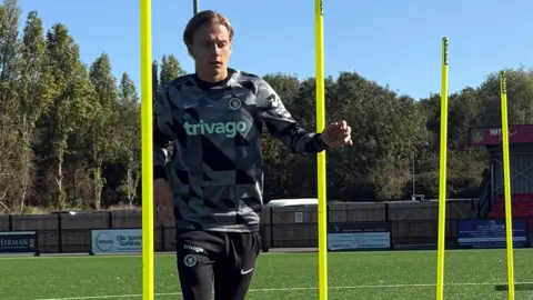 يرتدي جورج إدواردز قميصًا منقوشًا باللونين الرمادي والأسود وبنطال رياضي أسود من Nike. إنه يراوغ كرة القدم في ملعب خارجي. 