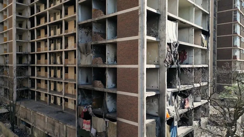 لقطة من طائرة بدون طيار لجانب مبنى مهجور. الواجهة مغطاة بالقماش مما يوفر الحماية لبعض من يعيشون بالداخل.