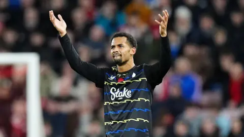 يرتدي PA Media Iliman Ndiaye قميصًا أسود مع خطوط متعرجة باللونين الأزرق والأصفر. رفع ذراعيه احتفالاً بعد تسجيله هدفاً.