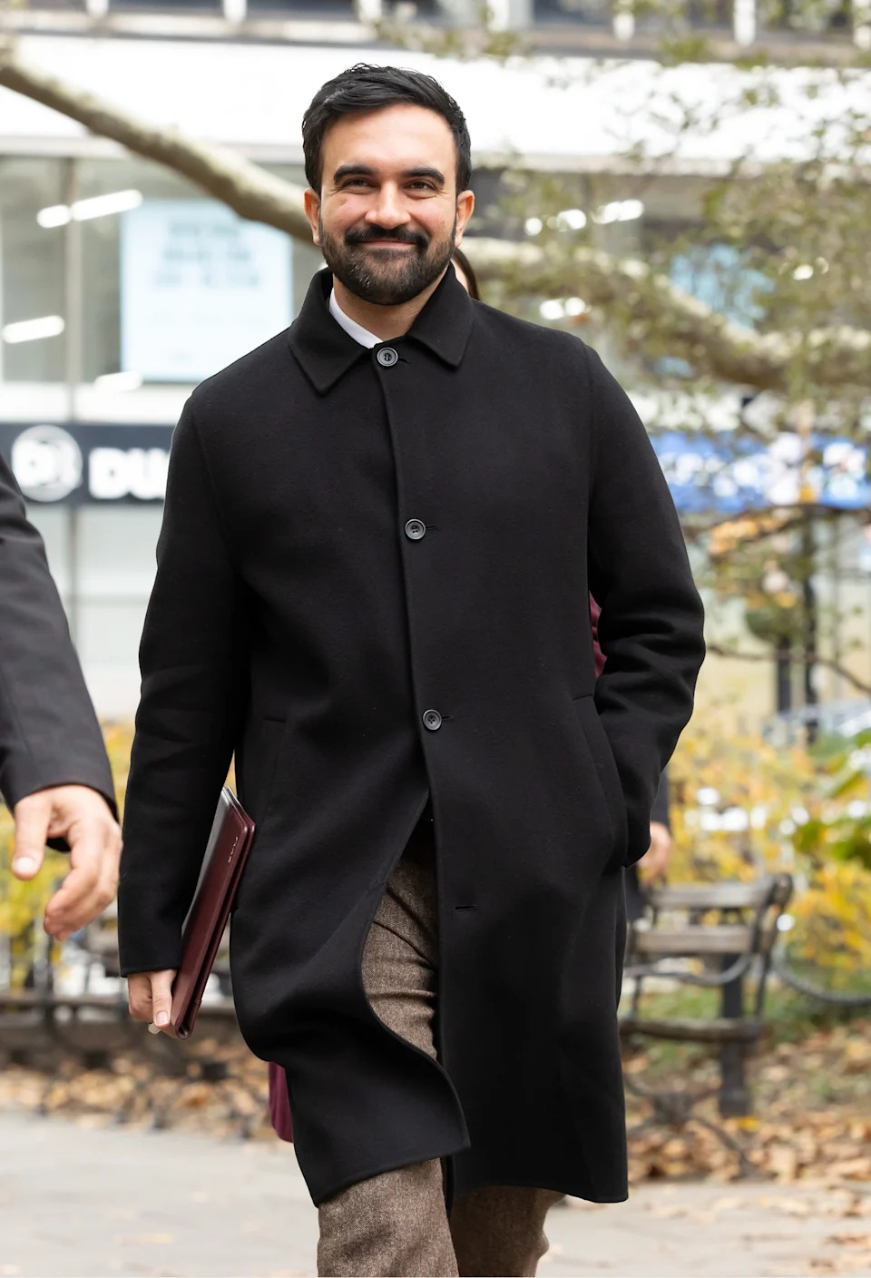 شخص يرتدي معطفًا بأزرار ويمشي في الخارج ويحمل مجلدًا، وتظهر الأشجار ومبنى في الخلفية