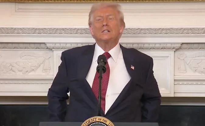 شخص يقف على منصة مع ختم، يرتدي بدلة وربطة عنق حمراء، في غرفة ذات مظهر رسمي مع مدفأة مزخرفة