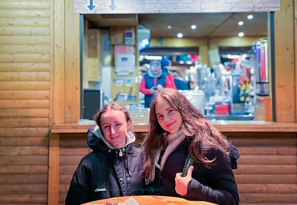 شابتان تجلسان على طاولة خشبية أمام كوخ خشبي تبيعان الفراولة المغطاة بالشوكولاتة في سوق عيد الميلاد في برلين. كلاهما يبتسم.