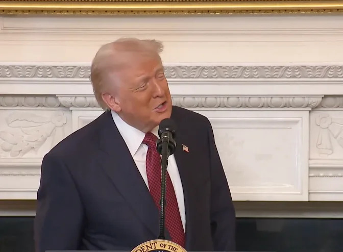 شخص يرتدي بدلة يتحدث على منصة بها مدفأة مزخرفة في الخلفية