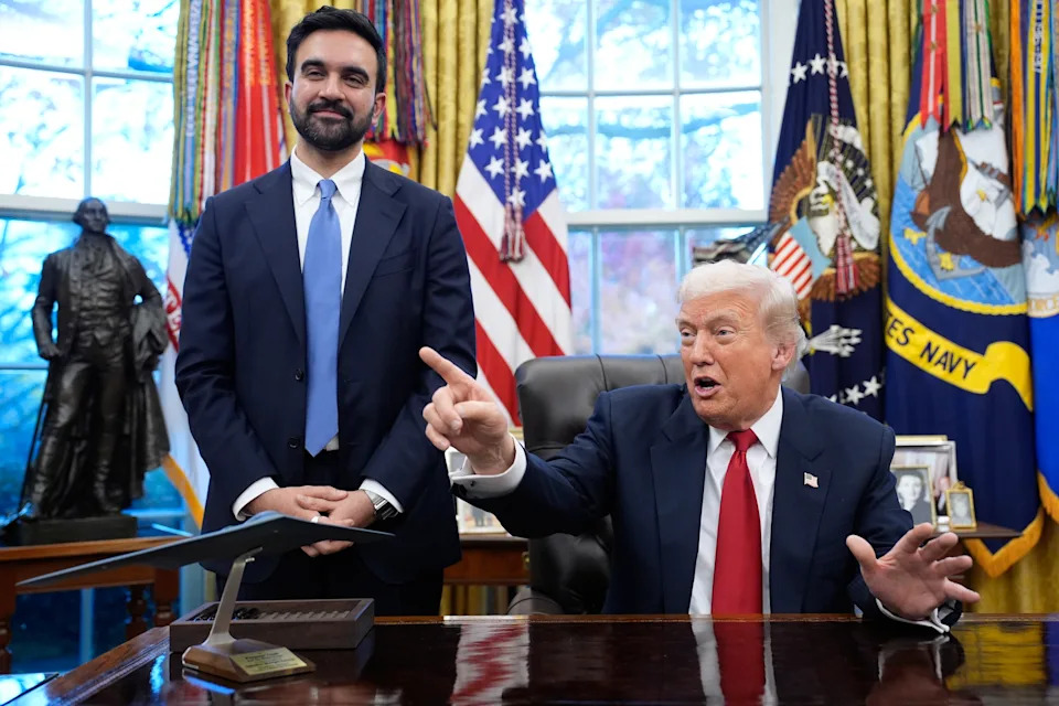 رجلان في أحد المكاتب. يشير الرجل الجالس أثناء التحدث. توجد أعلام مختلفة وتمثال في الخلفية