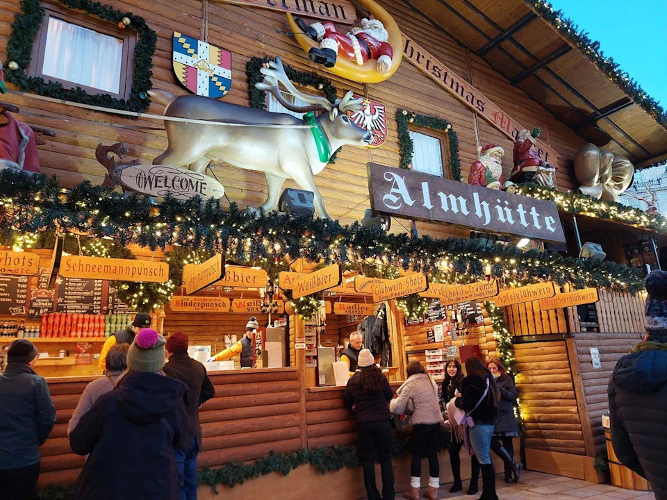 كشك لبيع الكحول في سوق عيد الميلاد. تم تزيين الكشك الخشبي بأغصان شجرة عيد الميلاد المزيفة، وأضواء خرافية ذهبية، وزخارف تشمل حيوان الرنة العملاق وزخارف سانتا، ولافتات تقول "المحوت", "فايسبير", "شنيمانبونش". تقف مجموعة من الأشخاص يرتدون معاطف شتوية دافئة وقبعات صوفية أمام الكشك، ويقدم لهم الموظفون الخدمة