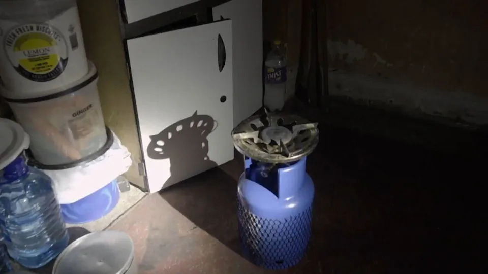 يتم إضاءة علبة غاز زرقاء وموقد بواسطة ضوء المشاعل. ويمكن رؤية زجاجة ماء وبعض الحاويات الأخرى في مكان قريب.