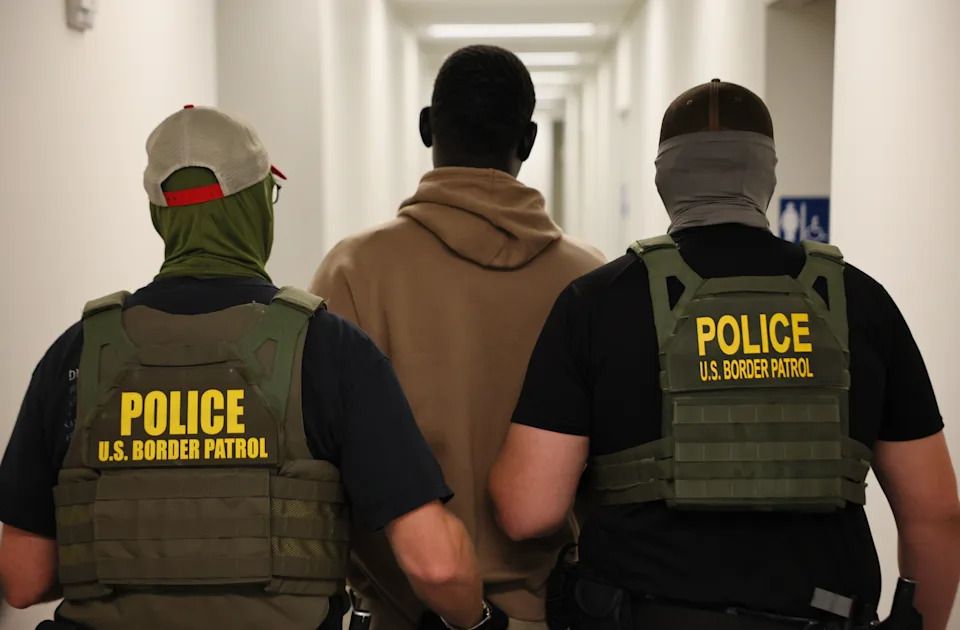 اثنان من ضباط حرس الحدود الأمريكيين يرافقان شخصًا في الردهة، ويرتدي معدات تكتيكية تحمل علامة "دورية الشرطة الحدودية الأمريكية."
