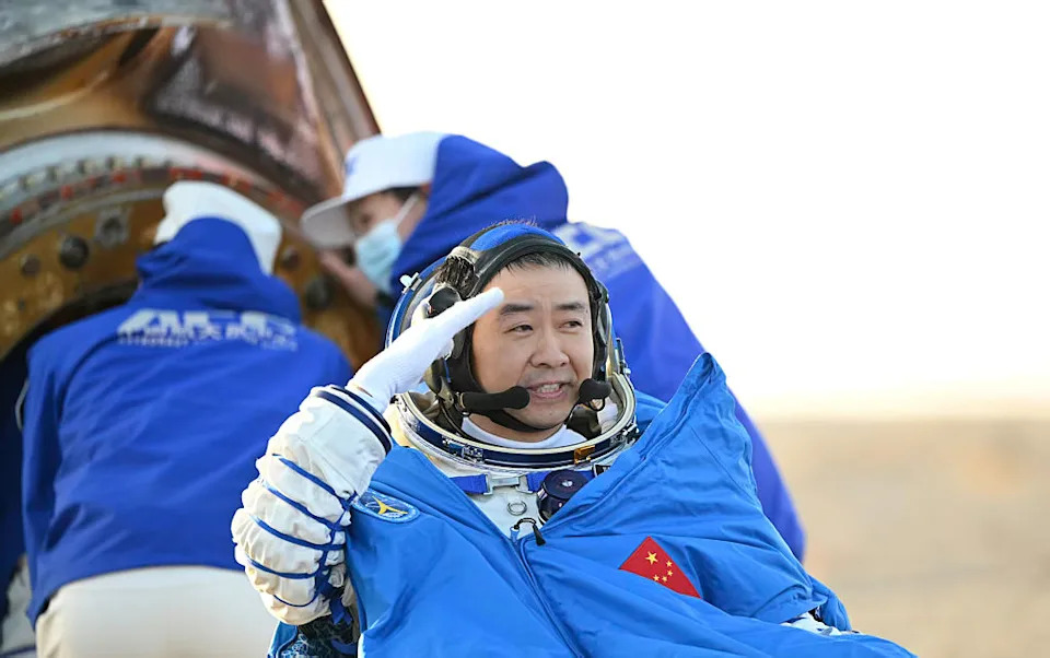 رجل يرتدي بدلة فضاء بيضاء وخوذة مفتوحة مع عباءة زرقاء فوق معداته مع علم أحمر بنجوم صفراء يحيي شخصًا خارج الكاميرا على يمين الصورة.