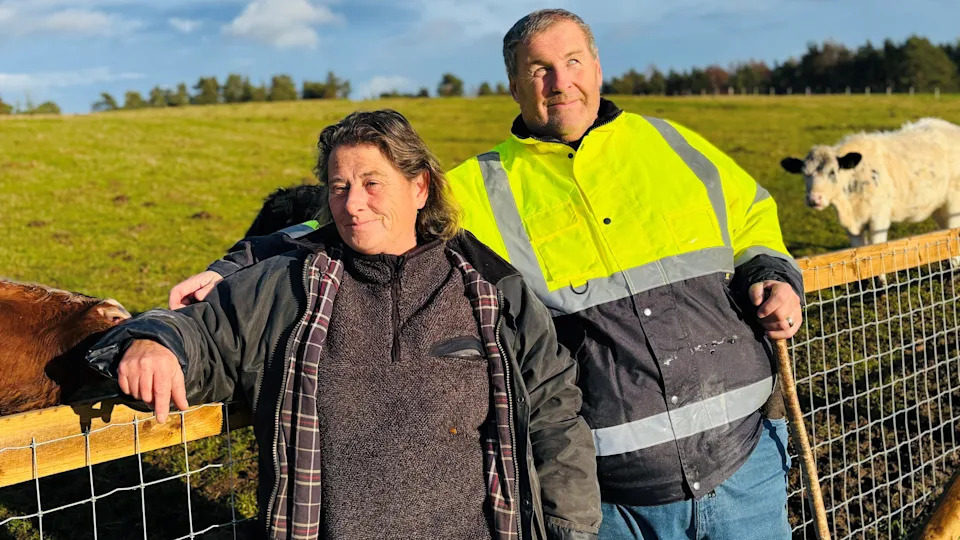 نيس ومايك دوكسبوري يبتسمان للكاميرا، وهي ترتدي ملابس على الطراز الزراعي، وهو يرتدي سترة أمان صفراء زاهية، وهناك أبقار في حقل خلفهما.