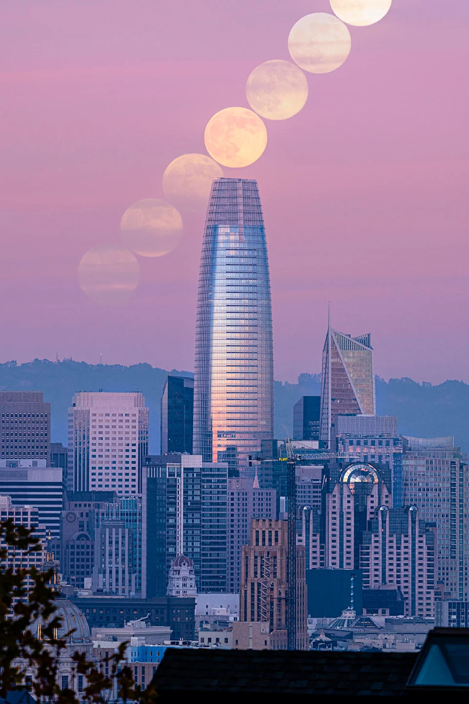 تشكل سلسلة من الأقمار الكاملة خطًا قطريًا في سماء المساء الوردية فوق مدينة حديثة مع ناطحة سحاب مركزية تهيمن على منتصف الصورة. 