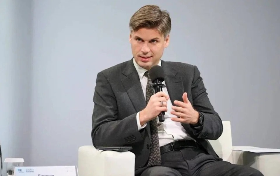 رجل ذو شعر رمادي يرتدي بدلة سوداء وبنطلون وقميص أبيض بأزرار، يجلس على كرسي أبيض ويحمل ميكروفونًا