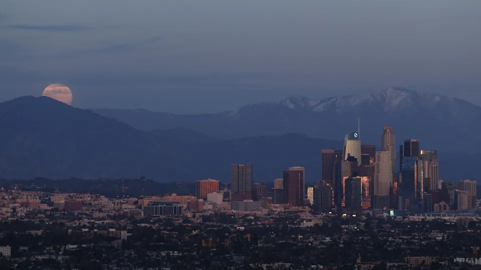 تم تصوير البدر وهو يرتفع فوق سلسلة جبال فوق إحدى المدن، بينما تتجمع ناطحات السحاب على يسار الصورة.