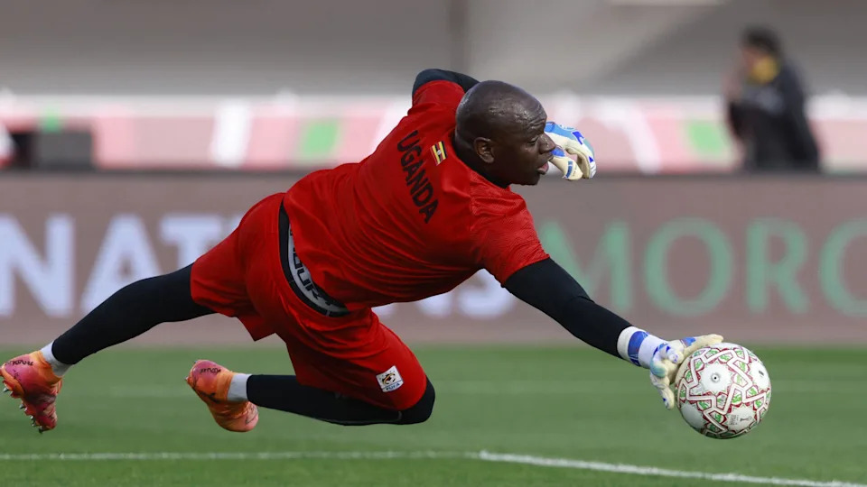 يقفز حارس المرمى دينيس أونيانجو إلى يمينه ليبعد الكرة أثناء عملية الإحماء قبل مباراة كأس الأمم الأفريقية 2025 ضد نيجيريا. يرتدي طقمًا أحمر اللون وطماقًا سوداء وأكمامًا تحتية وقفازات بيضاء. يحمل قميصه كلمة 