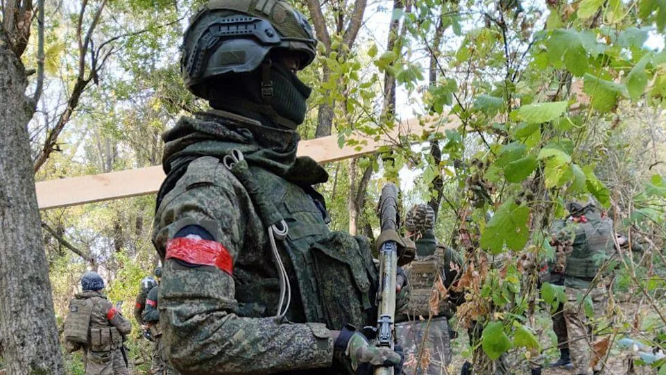 ديفيد كولوبا يرتدي معدات القتال الكاملة ويحمل مسدسًا في غابة في أوكرانيا مع جنود آخرين يظهرون في الخلفية.