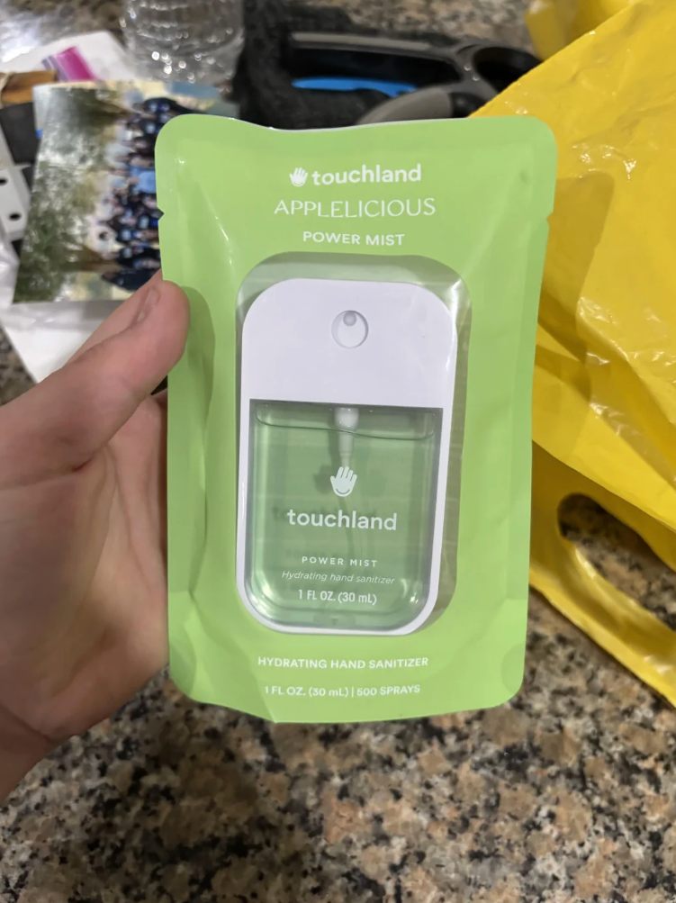 يد تحمل حزمة مكتوب عليها "تاتشلاند" بخلفية خضراء، تم شراؤها عن طريق الخطأ كعطر ولكنها تحتوي في الواقع على مطهر لليدين