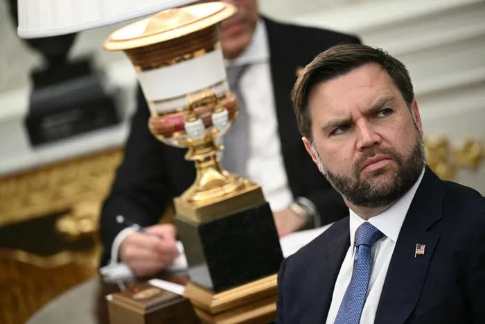 رجل يرتدي بدلة يجلس على طاولة الاجتماعات وينظر إلى الجانب. شخص في الخلفية يقوم بتدوين الملاحظات