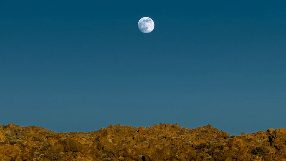 تم تصوير البدر وهو يرتفع فوق منظر طبيعي صخري في سماء زرقاء.