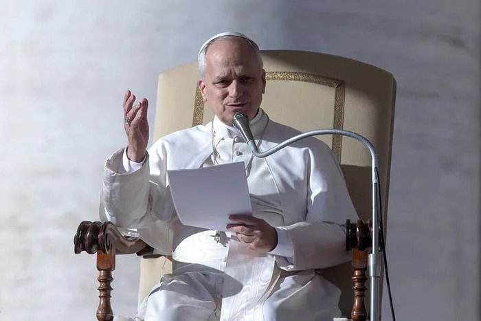 شخص يرتدي الزي البابوي يتحدث عبر الميكروفون بينما يحمل ورقة، ويجلس على كرسي كبير أثناء الحدث