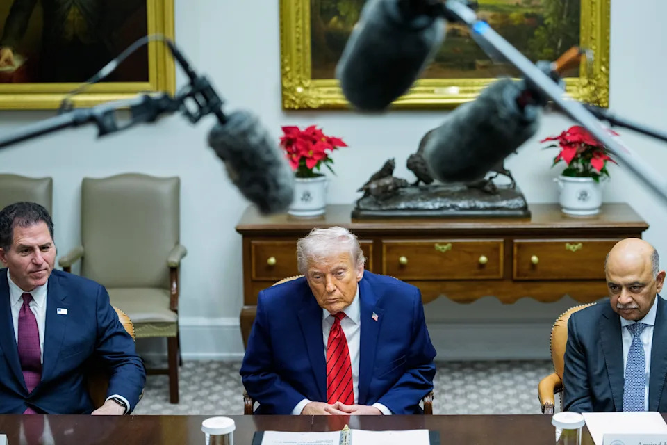 ثلاثة رجال يجلسون على طاولة اجتماعات مع وجود ميكروفونات في الأعلى. الرجل الموجود في المنتصف يرتدي بدلة زرقاء وربطة عنق حمراء. الإعداد رسمي