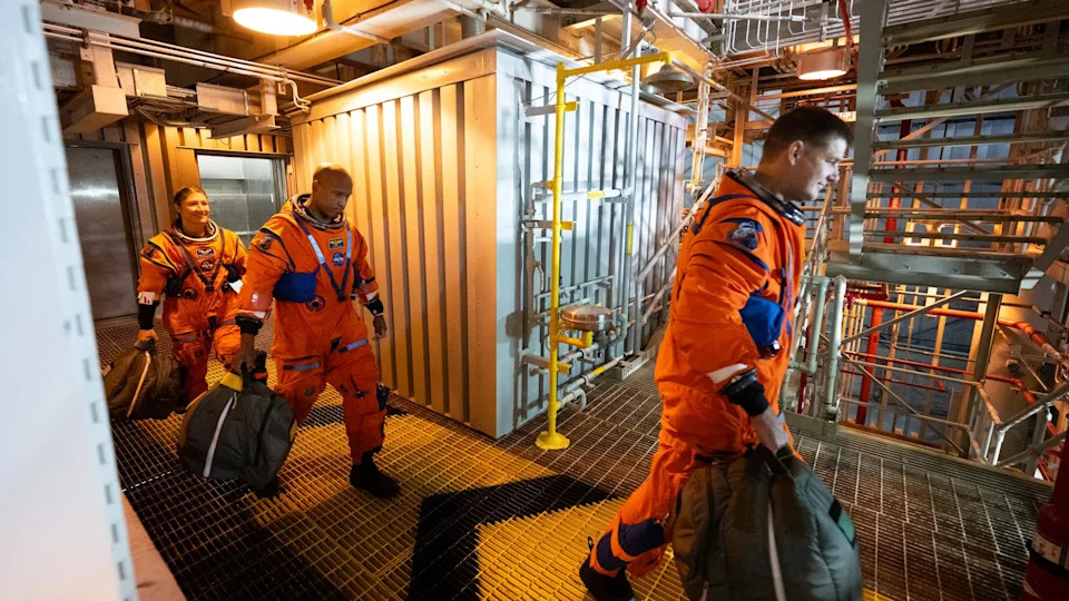 رائدا فضاء ورائدة فضاء، جميعهم يرتدون بدلات طيران برتقالية، يسيرون على جسر معدني داخل مبنى ضخم