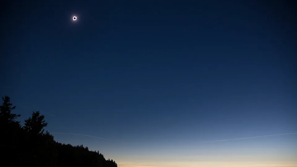 تظهر هالة كبيرة متوهجة لكسوف الشمس في سماء الليل الزرقاء الداكنة مع منظر طبيعي مظلل في مقدمة الصورة