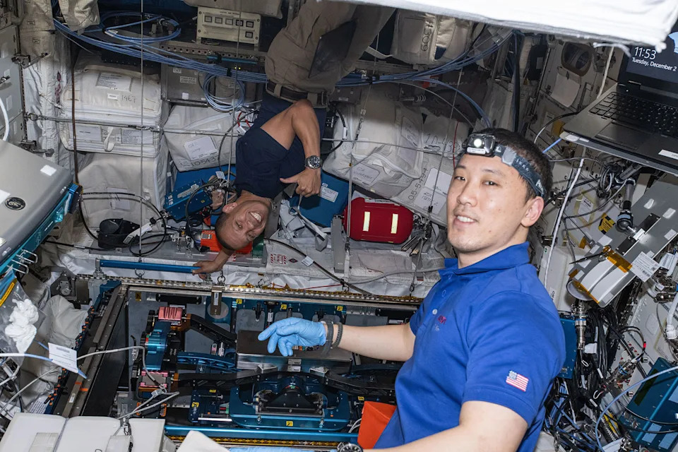 رجلان يرتديان قمصانًا زرقاء قصيرة الأكمام والآخر يرتدي قفازات سيليكون ومصباحًا رأسيًا، يعملان معًا لتركيب المعدات على متن محطة فضائية