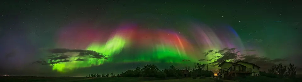 سلسلة من الشفق القطبي باللونين الأخضر والأحمر يمكن رؤيتها مع العديد من السحب والمنازل في صورة بانورامية كبيرة