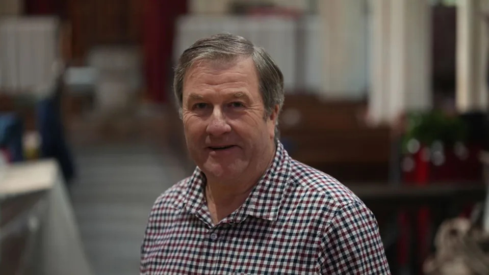 فيل جود يقف في الكنيسة. يرتدي قميصًا مربعًا وينظر إلى عدسة الكاميرا.